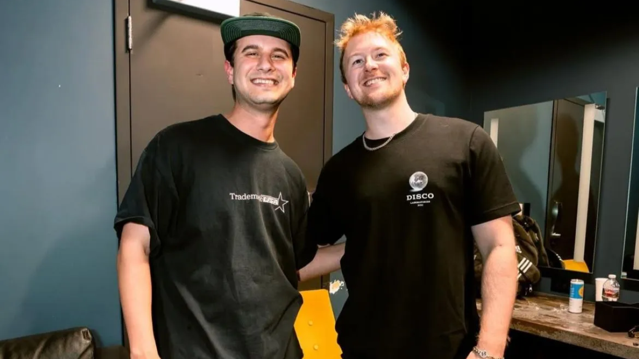 Tape B and Effin smiling together in a dressing room, both wearing black T-shirts, with mirrors and a counter visible behind them