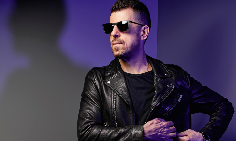 Sgt Slick wearing black sunglasses and a leather jacket in a purple-lit promotional portrait for his Strings Of Life remix