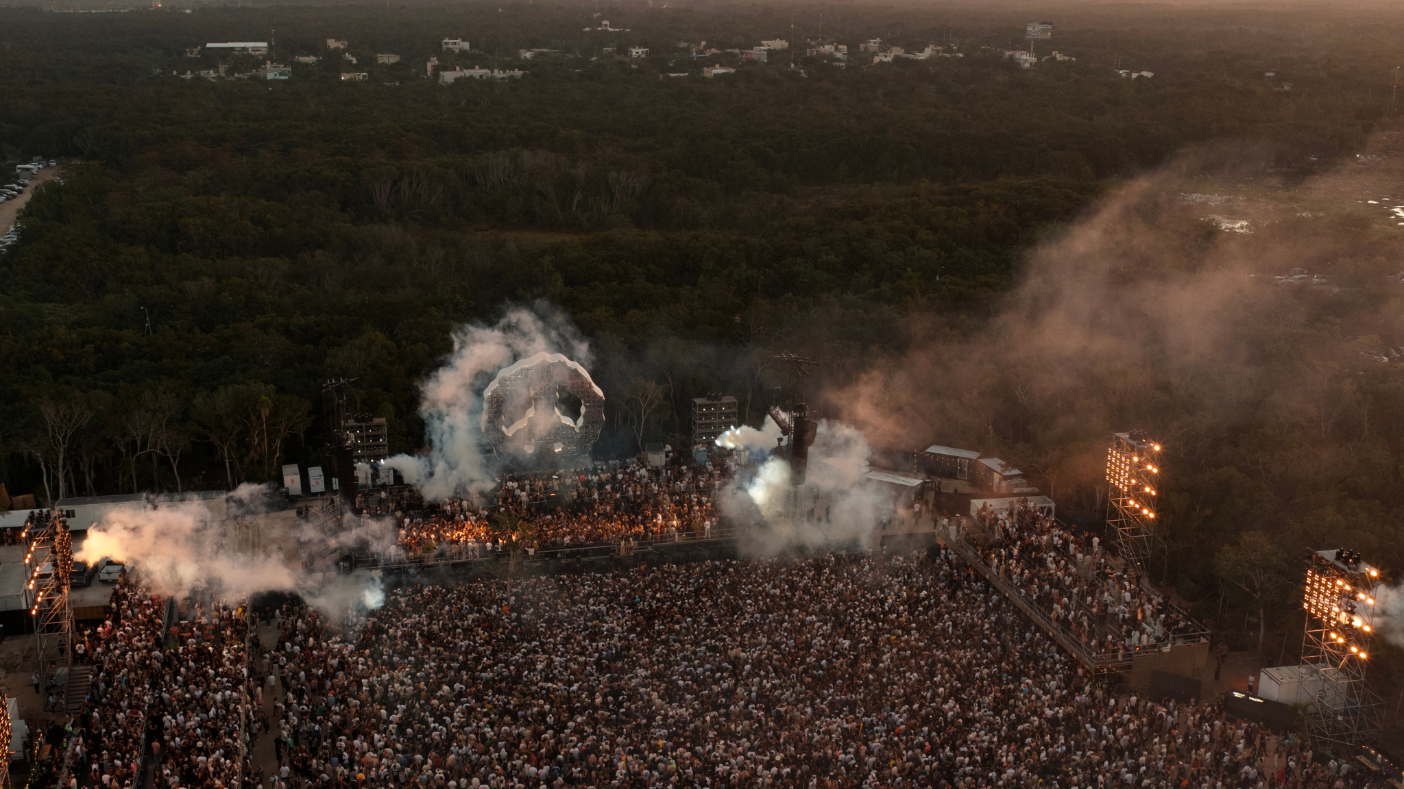 Aerial view of Zamna Tulum surrounded by jungle, with a large crowd, stage production, smoke and lighting at sunset
