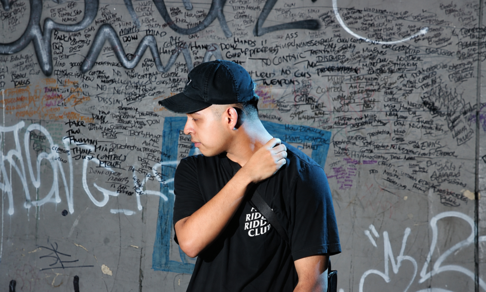 Deadlynoiz wearing a black cap and T-shirt in front of a graffiti-covered wall.