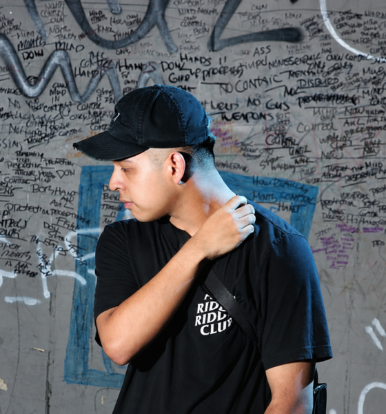 Deadlynoiz wearing a black cap and T-shirt in front of a graffiti-covered wall.