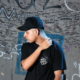 Deadlynoiz wearing a black cap and T-shirt in front of a graffiti-covered wall.