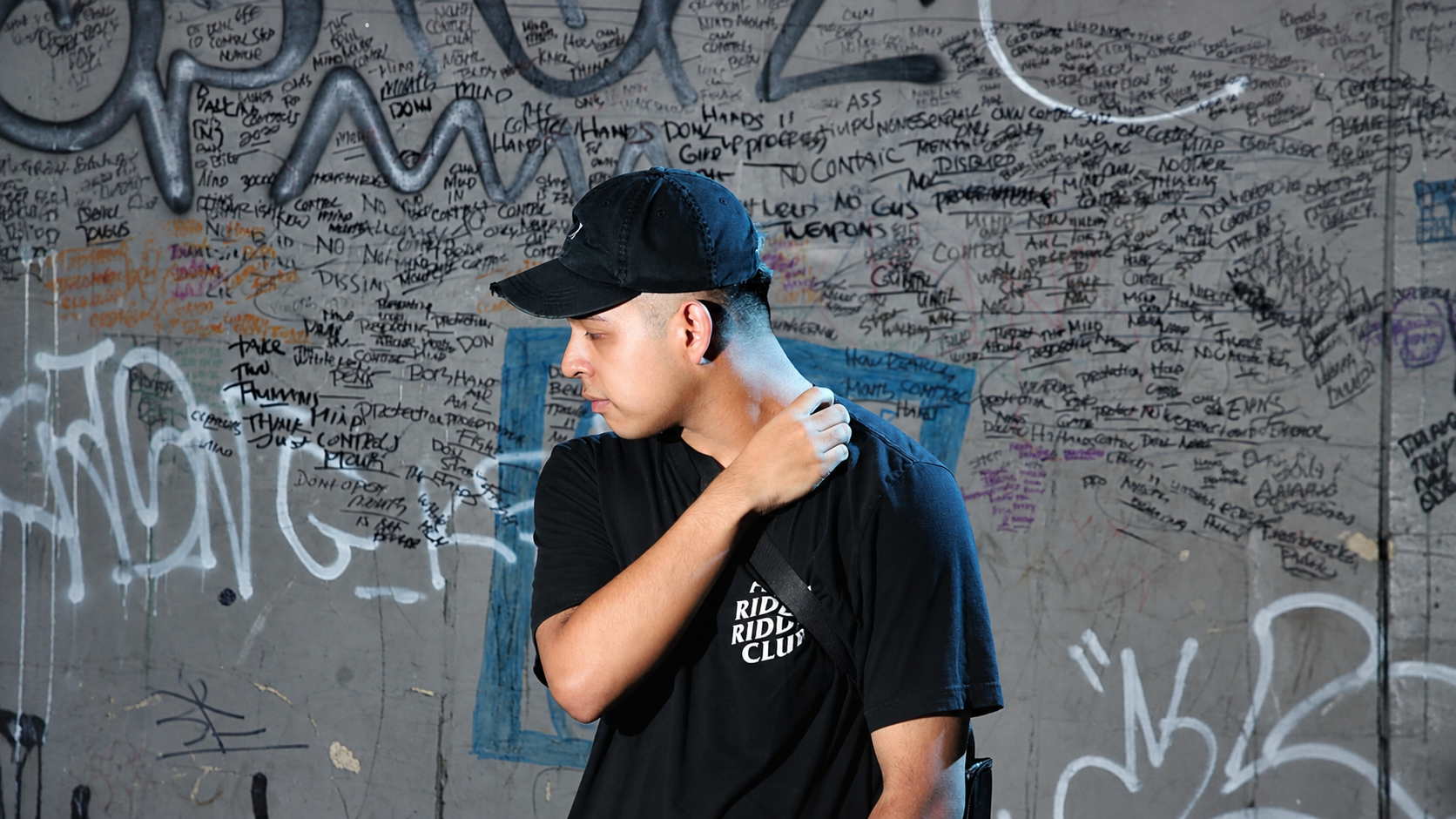 Deadlynoiz wearing a black cap and T-shirt in front of a graffiti-covered wall.
