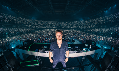 David Guetta standing behind his DJ decks in front of a crowd at a live electronic music performance.