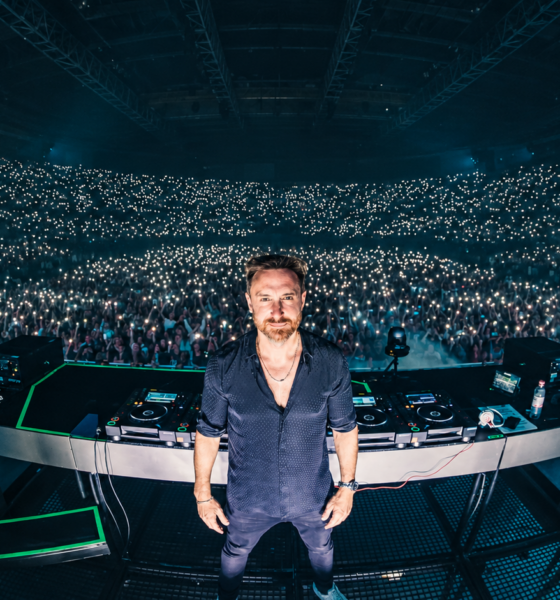 David Guetta standing behind his DJ decks in front of a crowd at a live electronic music performance.