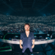 David Guetta standing behind his DJ decks in front of a crowd at a live electronic music performance.