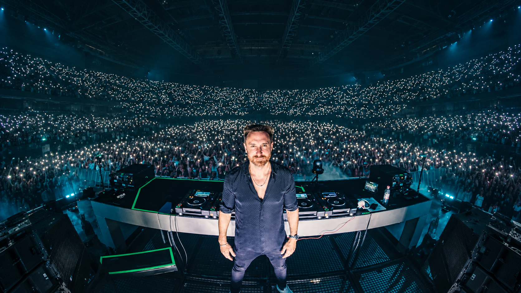 David Guetta standing behind his DJ decks in front of a crowd at a live electronic music performance.