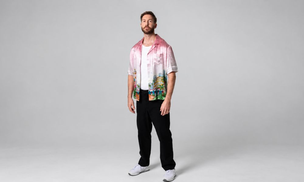Calvin Harris standing in a pink printed shirt, white T-shirt and black trousers against a grey studio backdrop