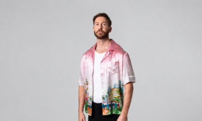 Calvin Harris standing in a pink printed shirt, white T-shirt and black trousers against a grey studio backdrop