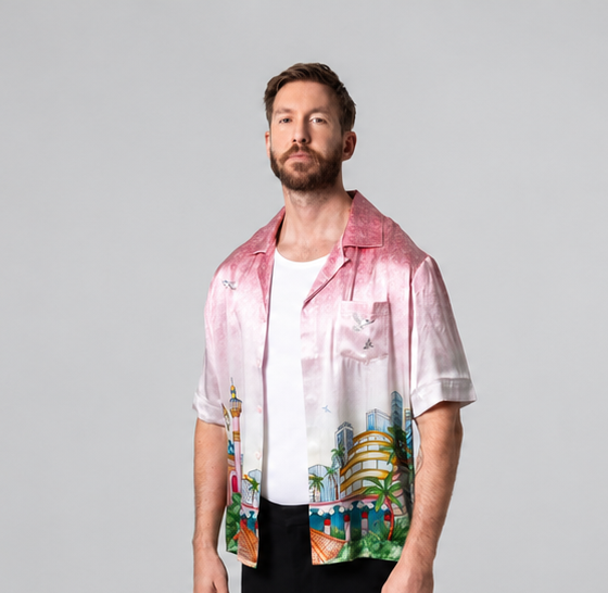 Calvin Harris standing in a pink printed shirt, white T-shirt and black trousers against a grey studio backdrop
