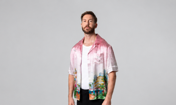 Calvin Harris standing in a pink printed shirt, white T-shirt and black trousers against a grey studio backdrop