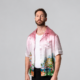 Calvin Harris standing in a pink printed shirt, white T-shirt and black trousers against a grey studio backdrop