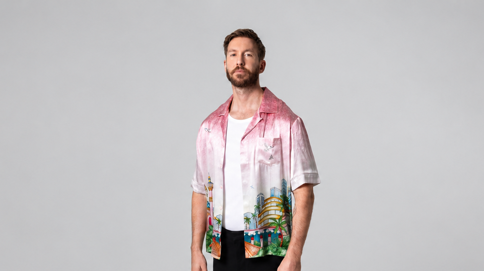 Calvin Harris standing in a pink printed shirt, white T-shirt and black trousers against a grey studio backdrop