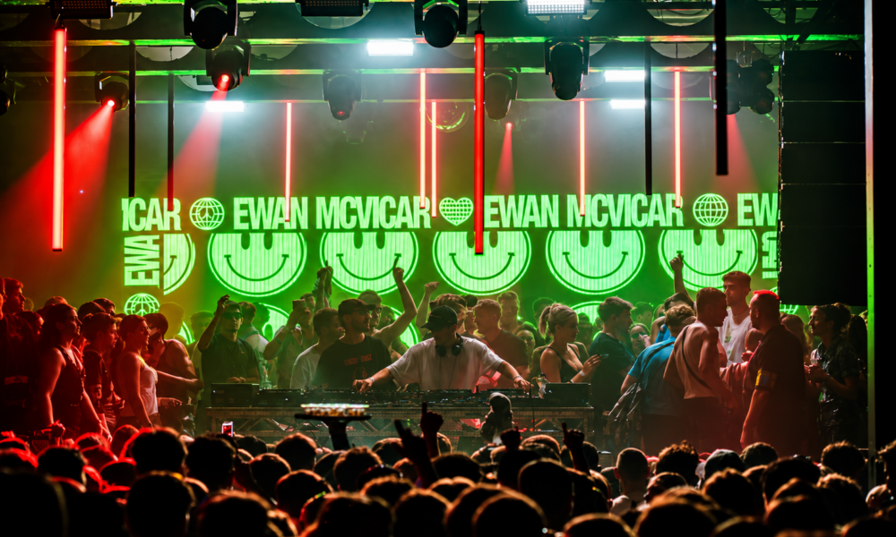Packed crowd in front of the DJ booth beneath green smiley-face screens and red strip lights