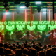 Packed crowd in front of the DJ booth beneath green smiley-face screens and red strip lights