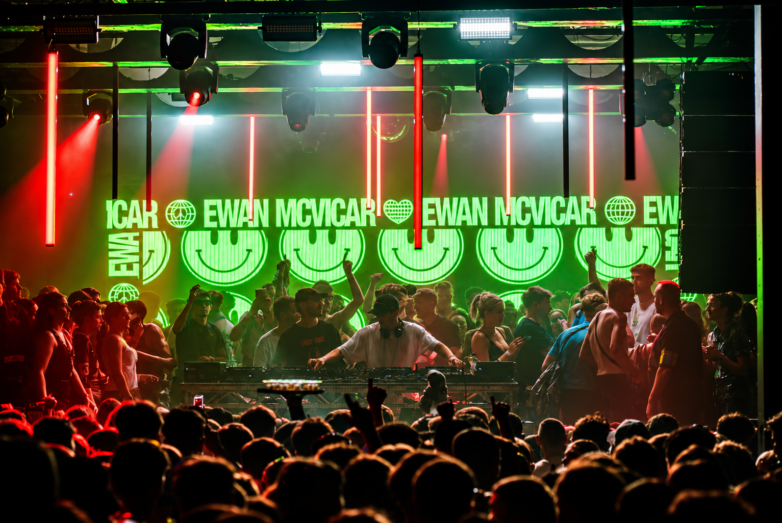 Packed crowd in front of the DJ booth beneath green smiley-face screens and red strip lights
