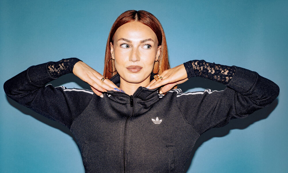 Scottish DJ and producer BETH posing with her hands under her chin in a black track jacket against a blue backdrop
