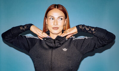 Scottish DJ and producer BETH posing with her hands under her chin in a black track jacket against a blue backdrop