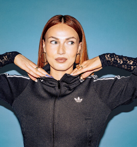 Scottish DJ and producer BETH posing with her hands under her chin in a black track jacket against a blue backdrop