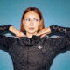 Scottish DJ and producer BETH posing with her hands under her chin in a black track jacket against a blue backdrop
