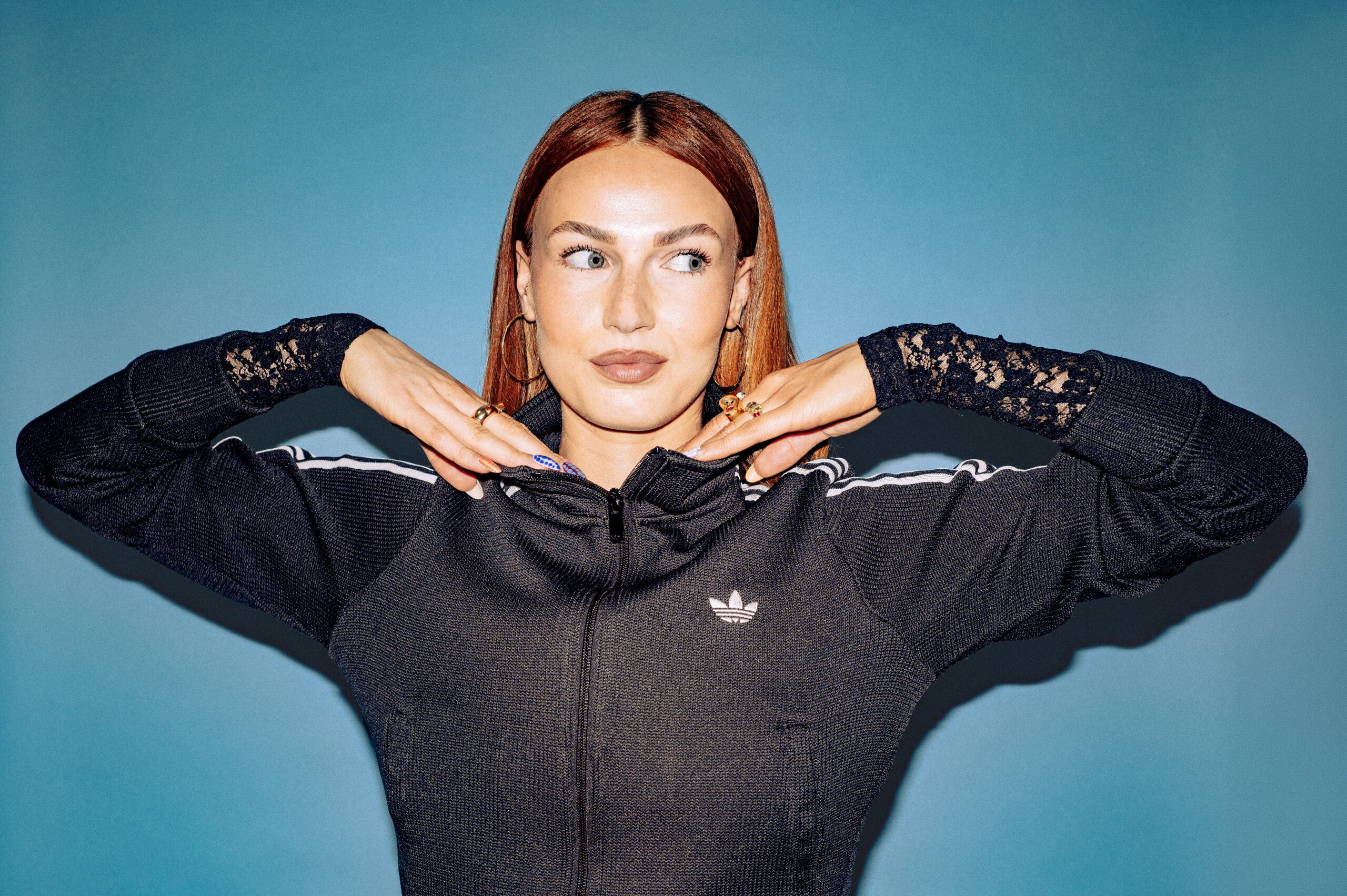 Scottish DJ and producer BETH posing with her hands under her chin in a black track jacket against a blue backdrop
