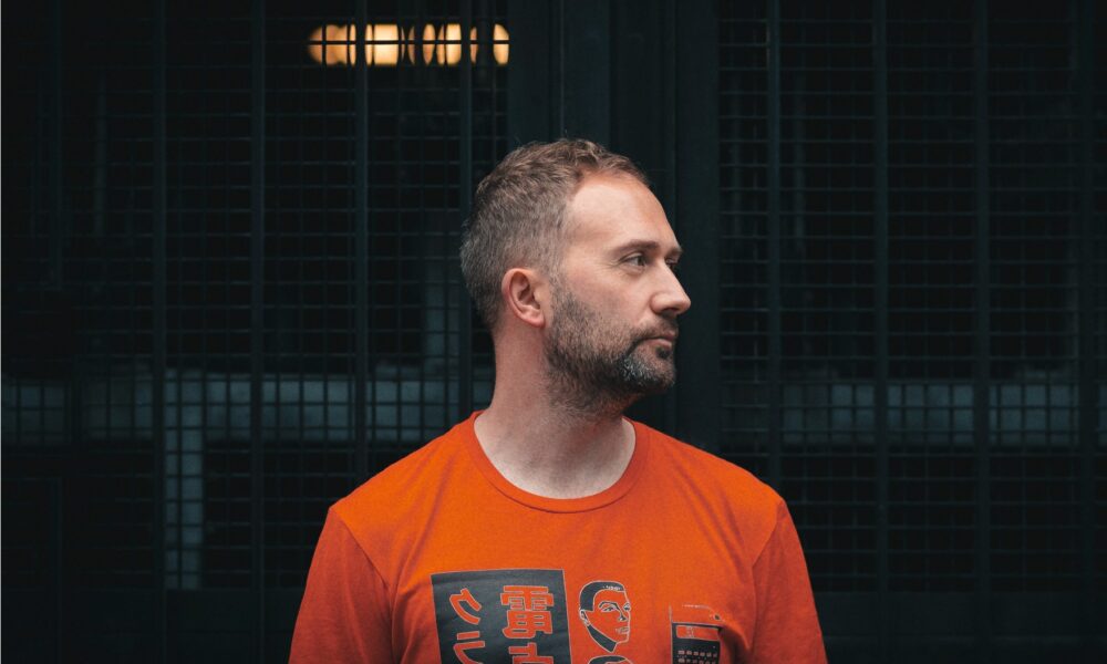 Scarper in an orange graphic T-shirt looking to the side in front of a dark metal grille