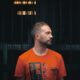 Scarper in an orange graphic T-shirt looking to the side in front of a dark metal grille