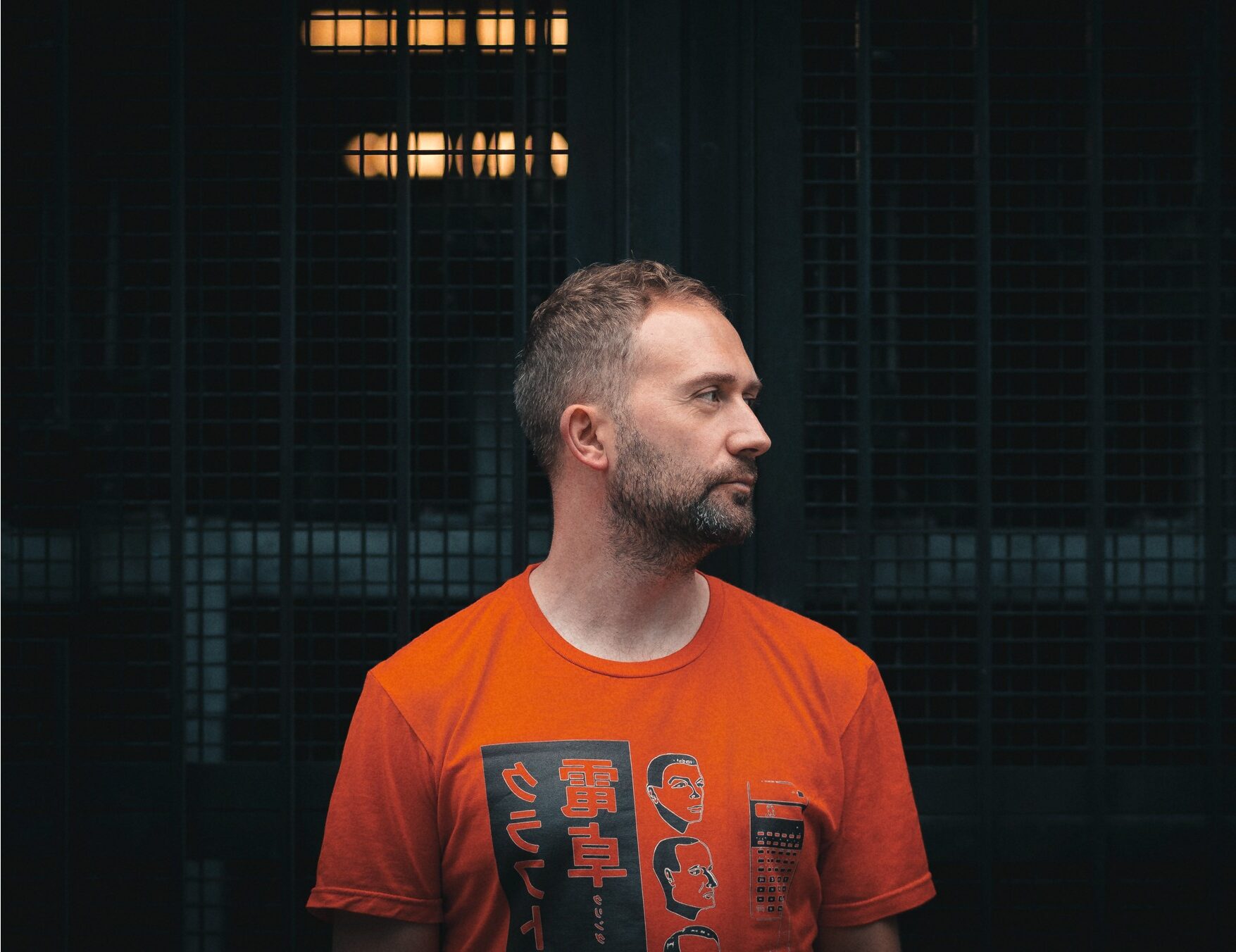 Scarper in an orange graphic T-shirt looking to the side in front of a dark metal grille