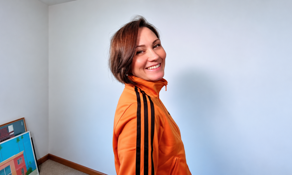 Edele Andaya smiling over her shoulder in an orange track jacket against a white wall