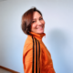 Edele Andaya smiling over her shoulder in an orange track jacket against a white wall
