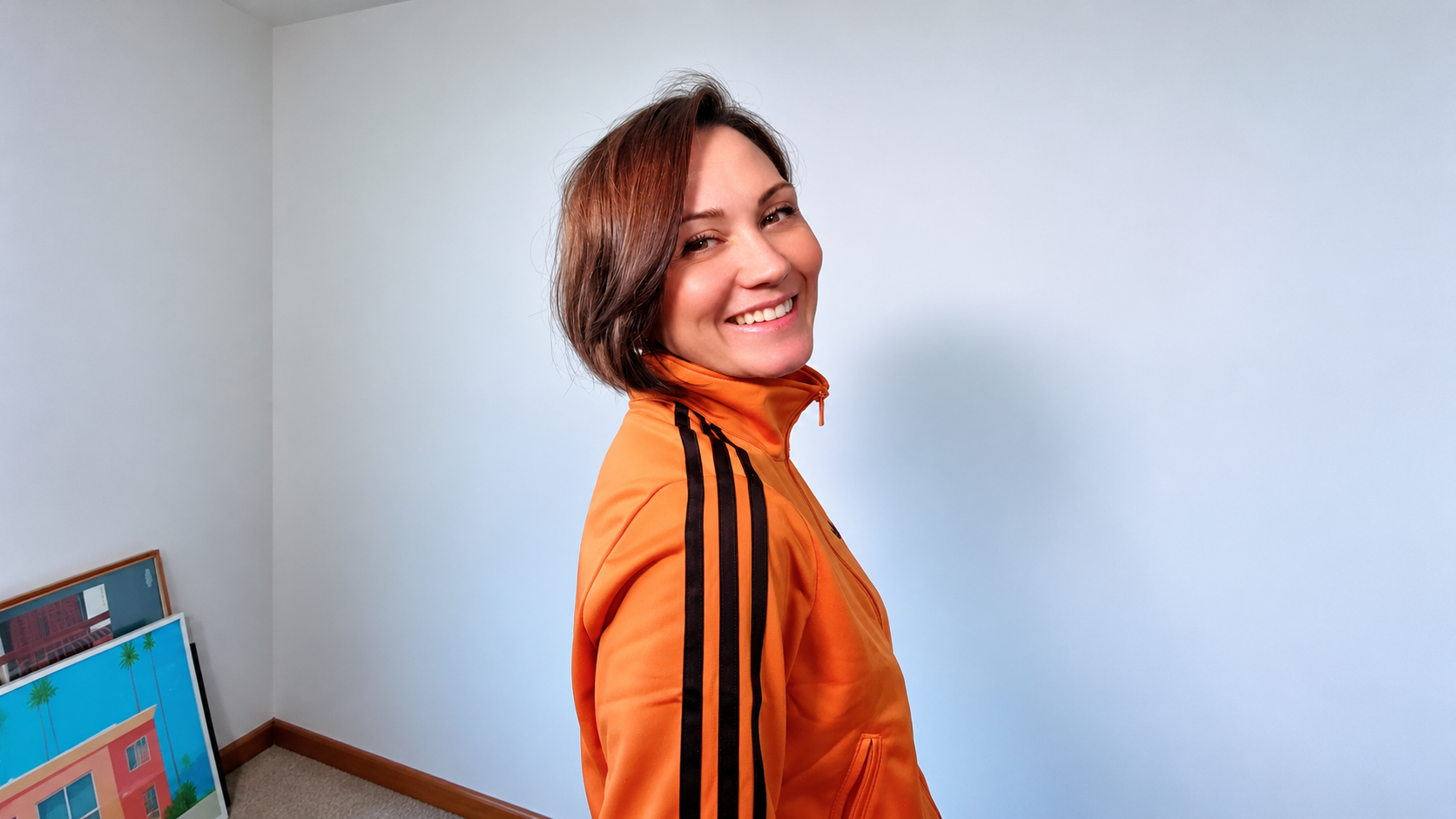Edele Andaya smiling over her shoulder in an orange track jacket against a white wall