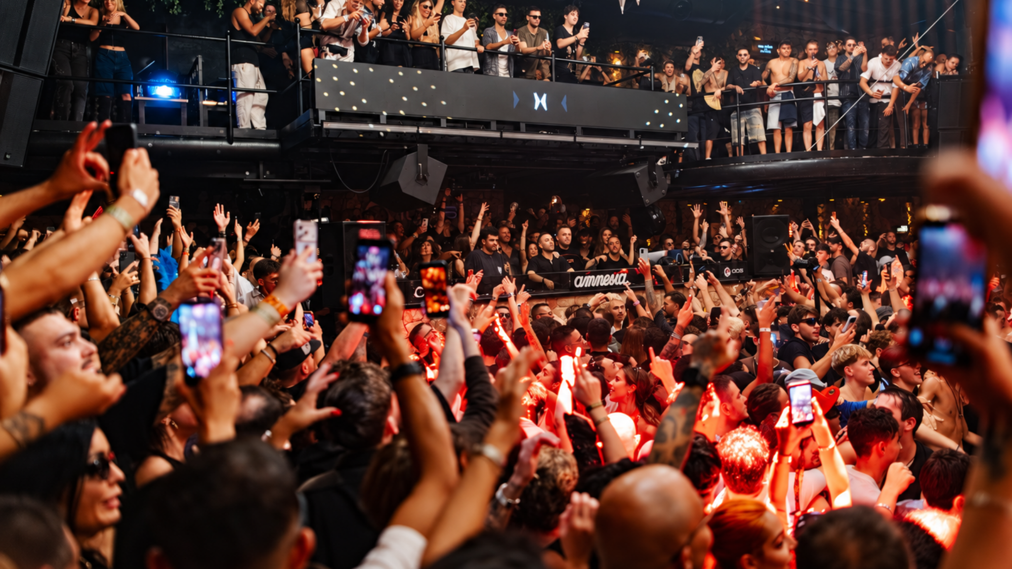 A crowded dancefloor at Amnesia Ibiza during the Metamorfosi 2026 closing, with the DJ booth surrounded by attendees under red lighting.