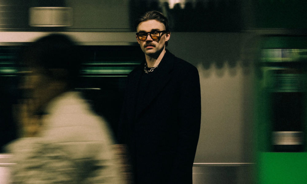 Dom Dolla wearing glasses and a black jacket in a blurred indoor setting