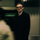 Dom Dolla wearing glasses and a black jacket in a blurred indoor setting