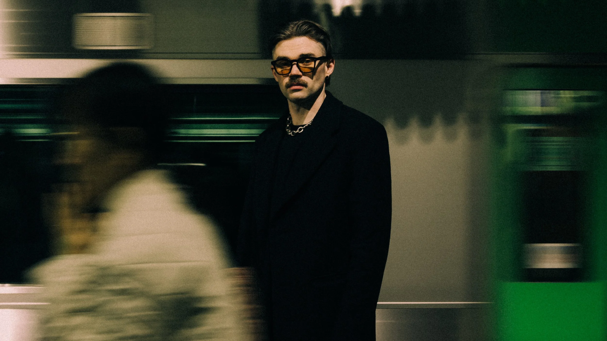 Dom Dolla wearing glasses and a black jacket in a blurred indoor setting