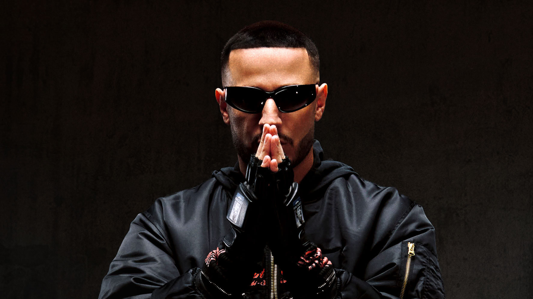 DJ Snake wearing black sunglasses and a black jacket with his hands together