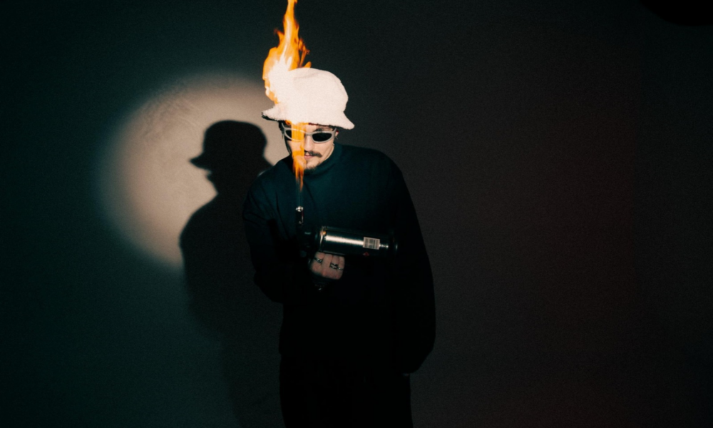 REYUS wearing sunglasses and a white hat with flames rising above it during a promotional shoot