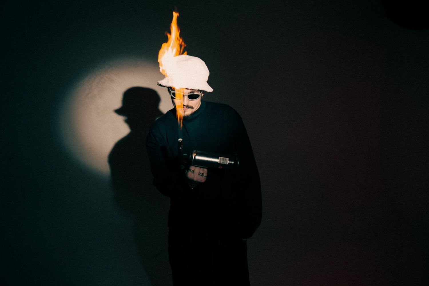 REYUS wearing sunglasses and a white hat with flames rising above it during a promotional shoot