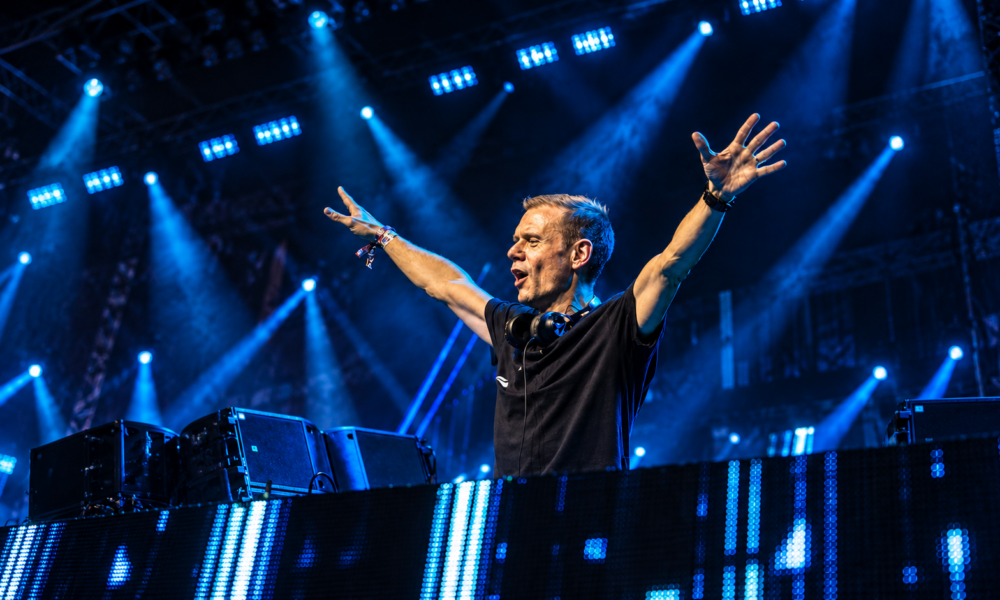 Armin van Buuren performing behind the DJ booth with his arms raised under blue stage lights, featured in New EDM Friday Oct 23
