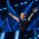 Armin van Buuren performing behind the DJ booth with his arms raised under blue stage lights, featured in New EDM Friday Oct 23