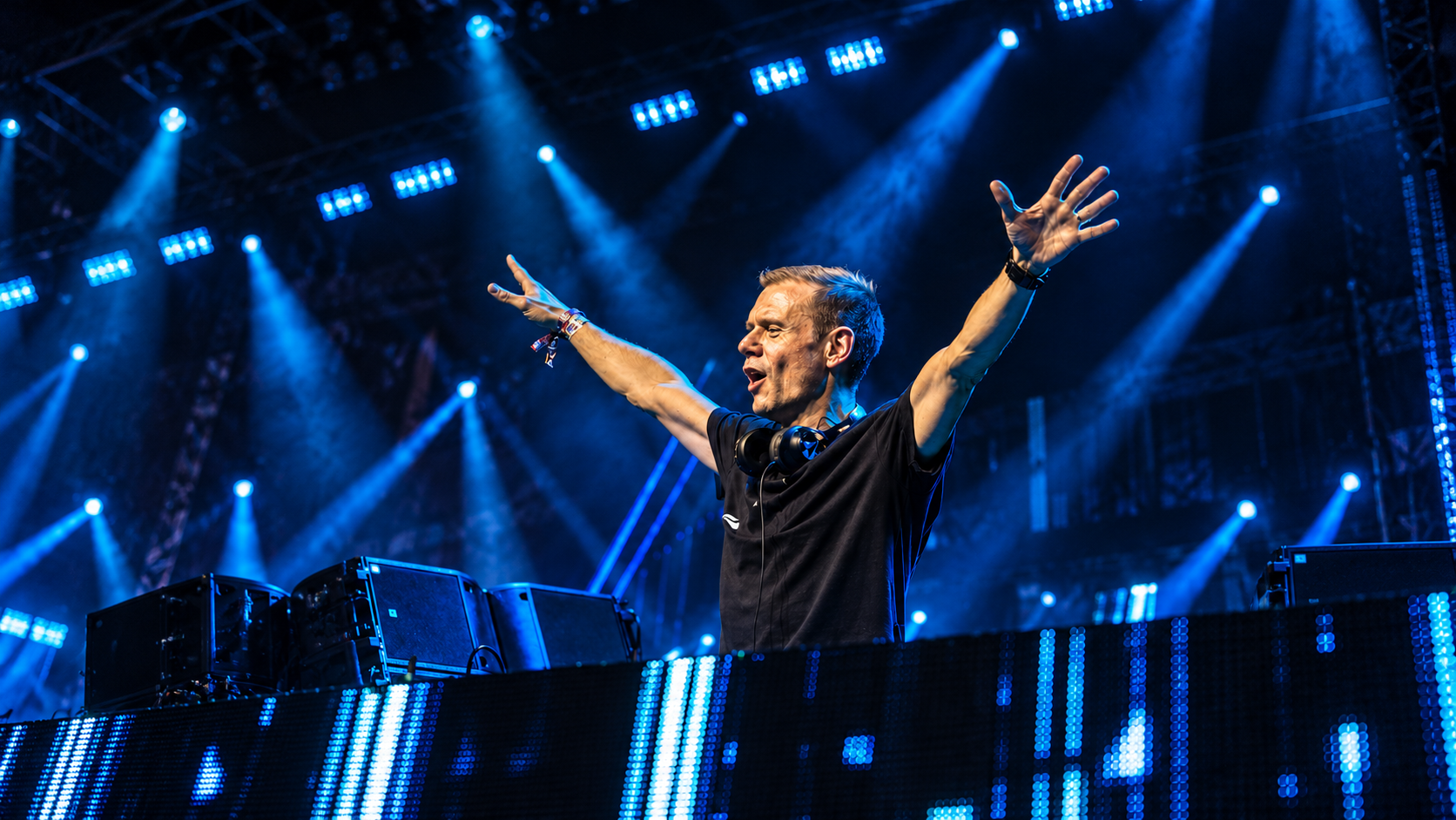 Armin van Buuren performing behind the DJ booth with his arms raised under blue stage lights, featured in New EDM Friday Oct 23