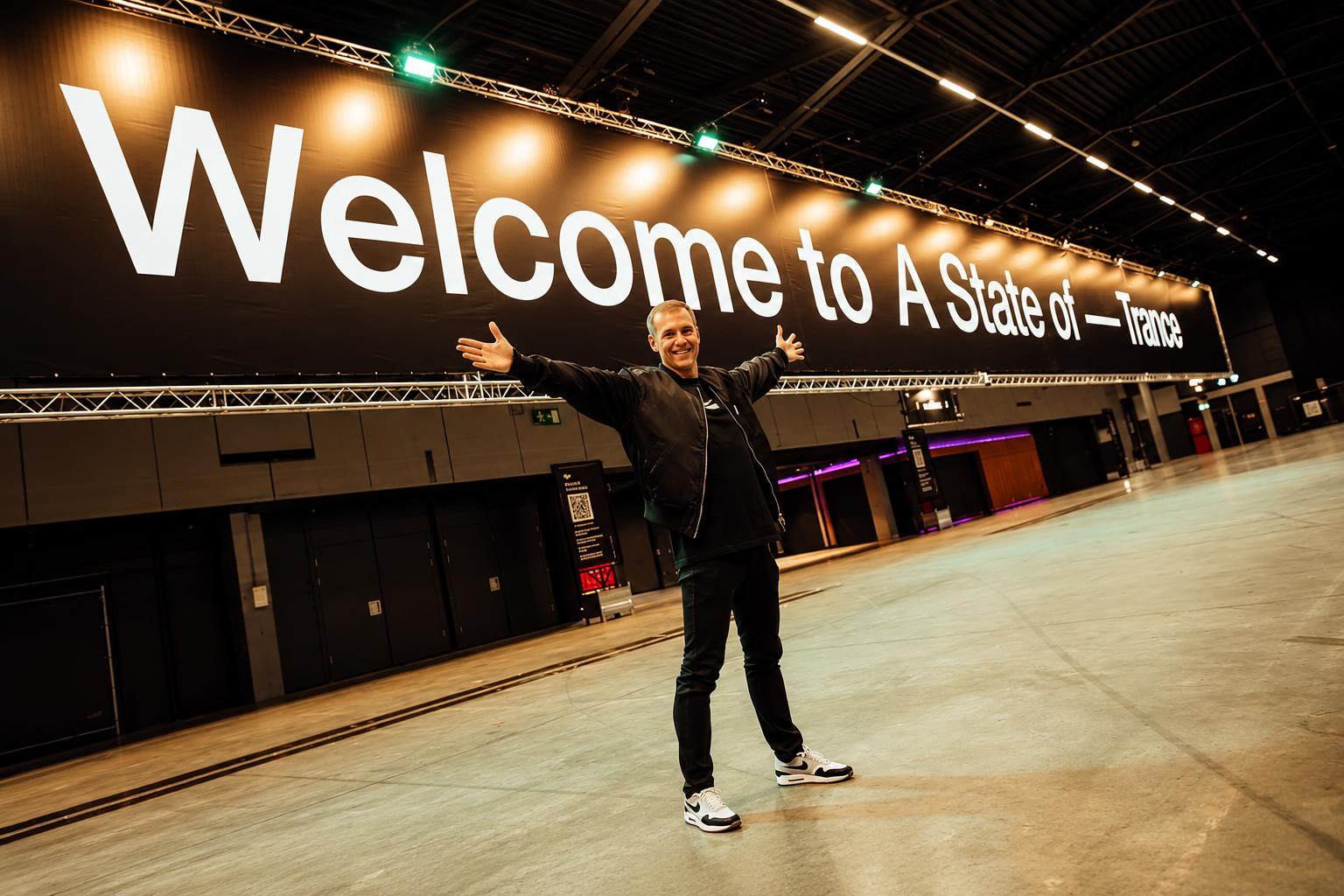 Armin van Buuren standing beneath a large “Welcome to A State of Trance” sign inside an event venue