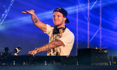 Avicii performing behind the DJ booth under blue stage lighting with one arm raised.