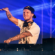 Avicii performing behind the DJ booth under blue stage lighting with one arm raised.
