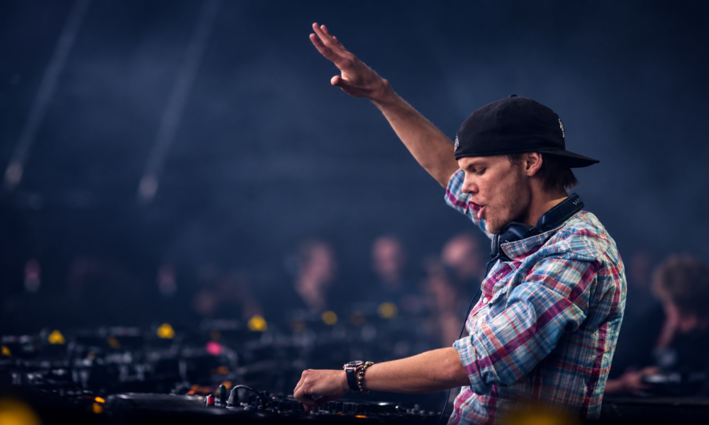 Avicii performing live behind the DJ decks with one arm raised, wearing a backwards black cap and plaid shirt during a festival performance.
