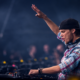 Avicii performing live behind the DJ decks with one arm raised, wearing a backwards black cap and plaid shirt during a festival performance.