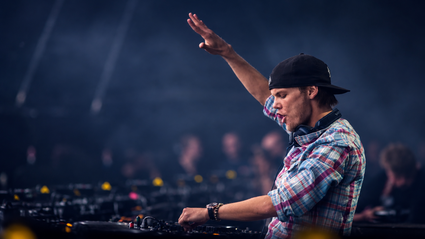 Avicii performing live behind the DJ decks with one arm raised, wearing a backwards black cap and plaid shirt during a festival performance.