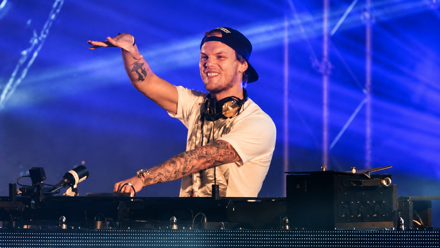 Avicii performing behind the DJ booth under blue stage lighting with one arm raised.
