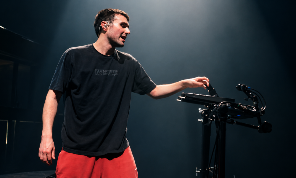Fred again.. performing live on stage beside his equipment under overhead lighting.