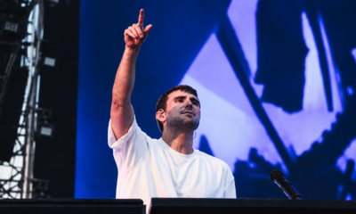 Fred again.. performing on stage with one arm raised in front of a blue screen